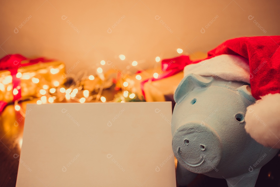 Cofrinho com chapéu de Papai Noel em fundo desfocado de Natal com enfeites, presentes e luz. Espaço para texto.