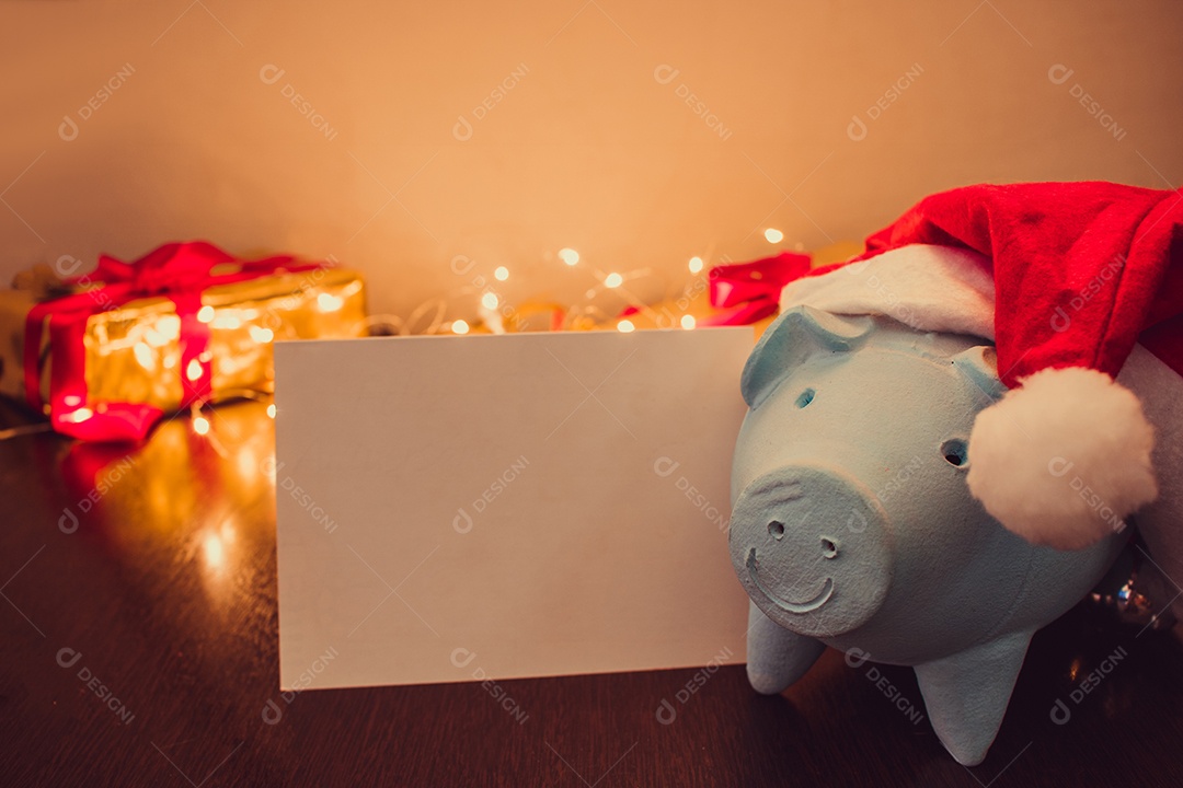Cofrinho com chapéu de Papai Noel em fundo desfocado de Natal com enfeites, presentes e luz. Espaço para texto.