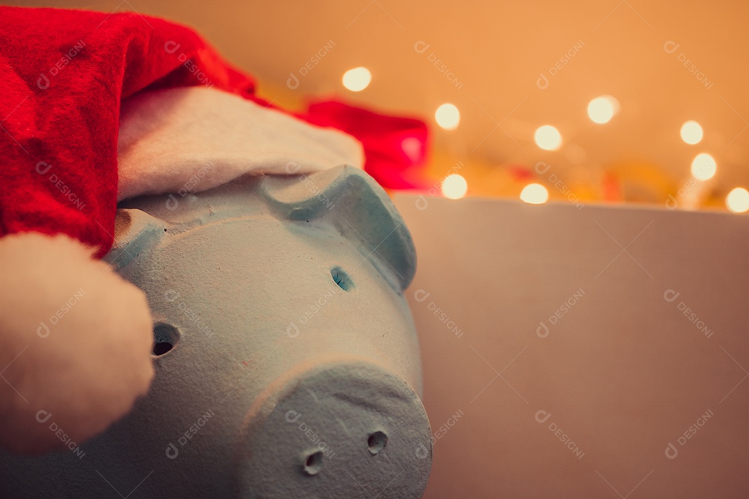 Cofrinho com chapéu de Papai Noel em fundo desfocado de Natal com enfeites, presentes e luz. Espaço para texto.