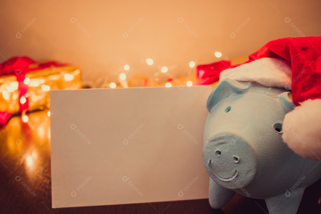 Cofrinho com chapéu de Papai Noel em fundo desfocado de Natal com enfeites, presentes e luz. Espaço para texto.