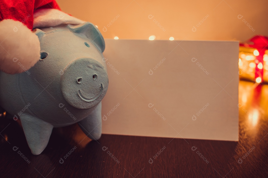 Cofrinho com chapéu de Papai Noel em fundo desfocado de Natal com enfeites, presentes e luz. Espaço para texto.