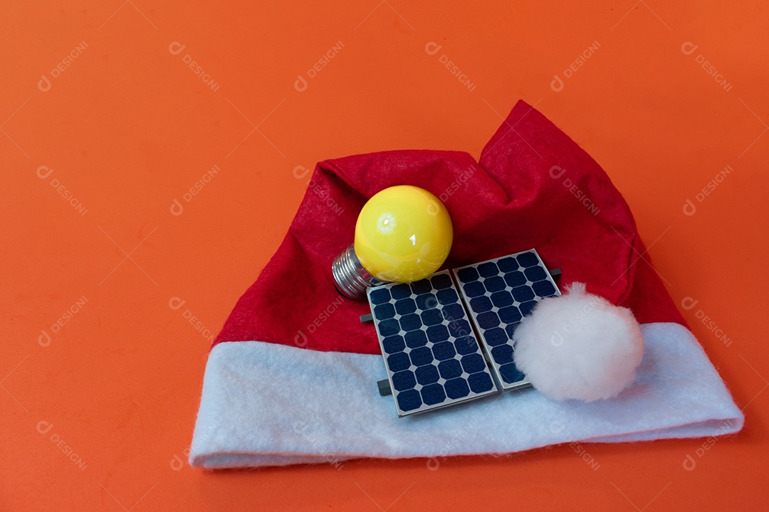 Chapéu de Papai Noel de Natal com painel solar fotovoltaico e lâmpada isolada em fundo laranja.