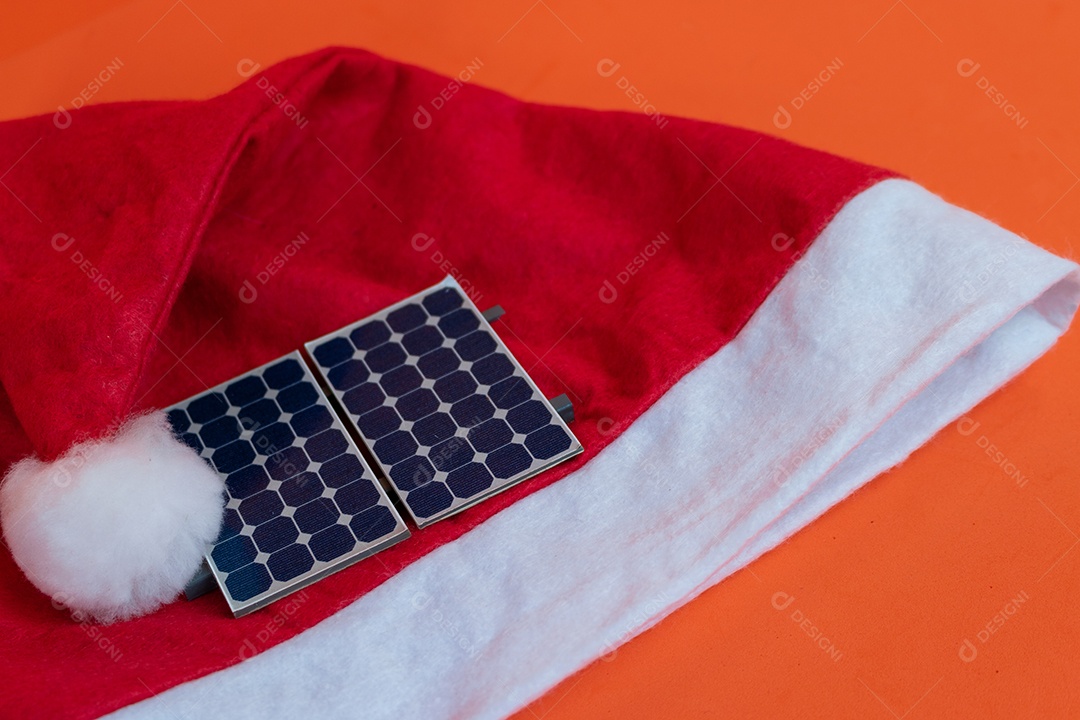 Chapéu de Papai Noel de Natal com painel solar fotovoltaico e lâmpada isolada em fundo laranja.