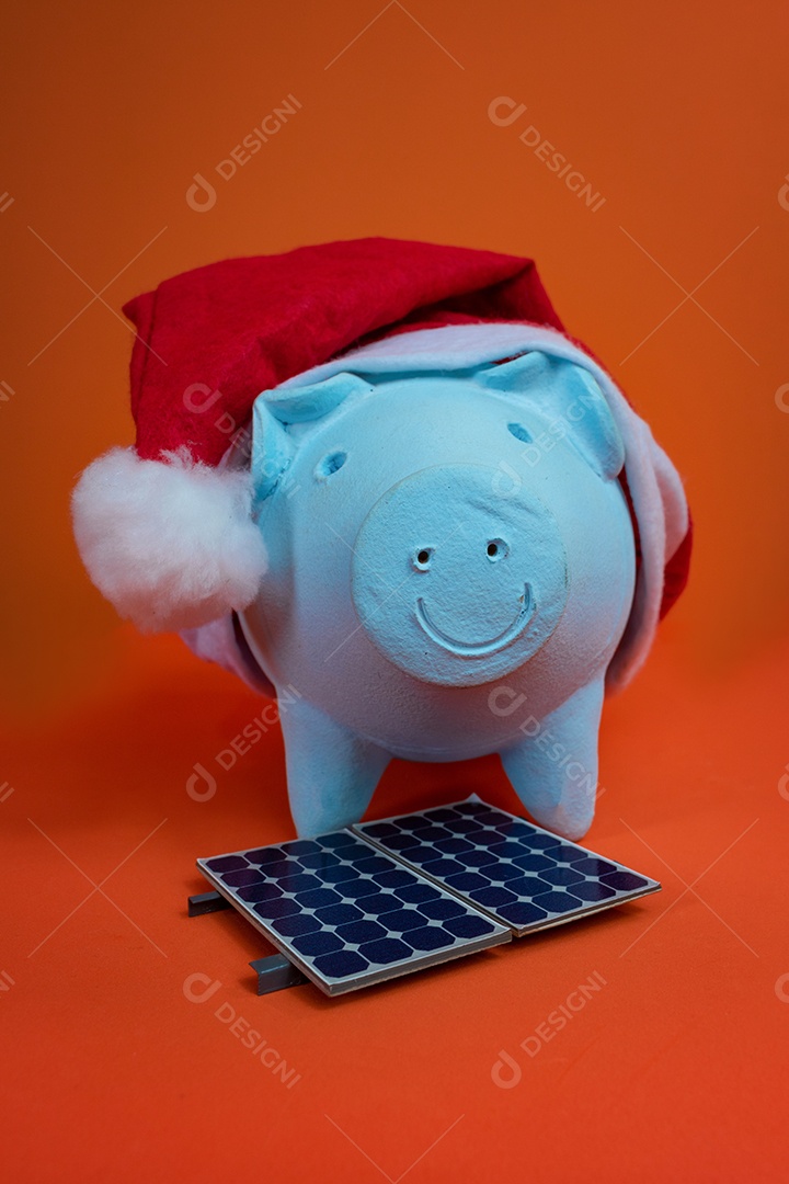 Moedas do cofrinho com chapéu de Natal de Papai Noel, painel solar fotovoltaico e lâmpada isolada em fundo laranja.