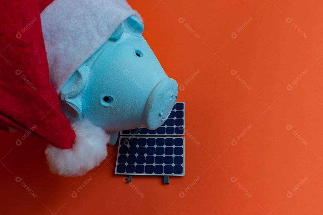 Moedas do cofrinho com chapéu de Natal de Papai Noel, painel solar fotovoltaico e lâmpada isolada em fundo laranja.