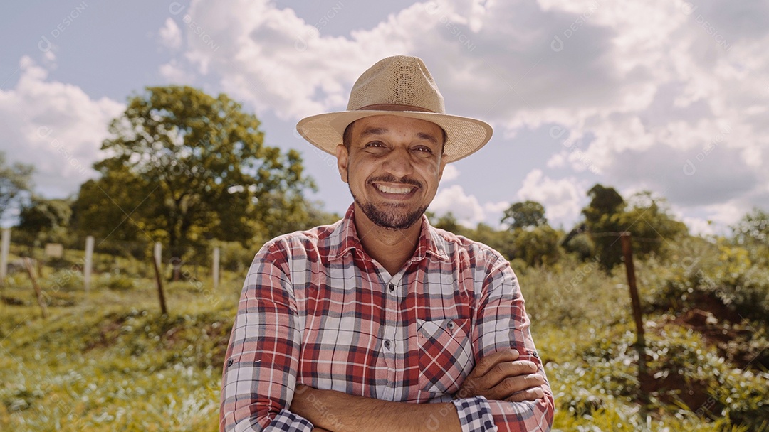 Retrato de jovem agricultor com mãos cruzadas na camisa casual e chapéu na fazenda