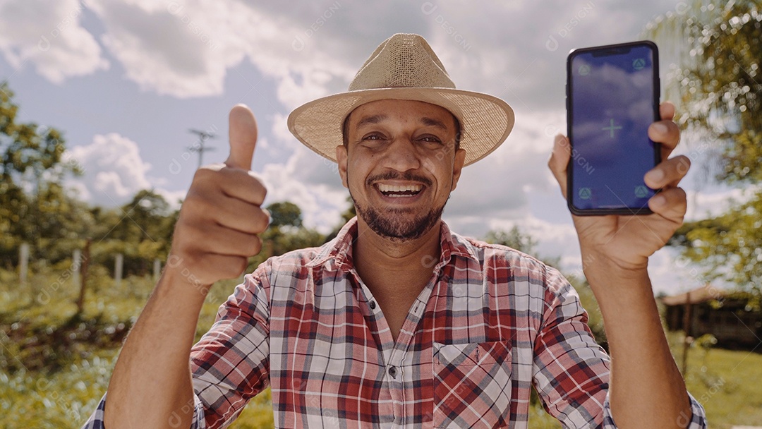 Jovem agricultor de chapéu sorrindo para a câmera e mostrando smartphone com tela de croma chave. Pastor latino feliz segurando o celular e demonstrando propaganda