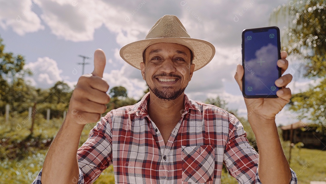 Jovem agricultor de chapéu sorrindo para a câmera e mostrando smartphone com tela de croma chave. Pastor latino feliz segurando o celular e demonstrando propaganda