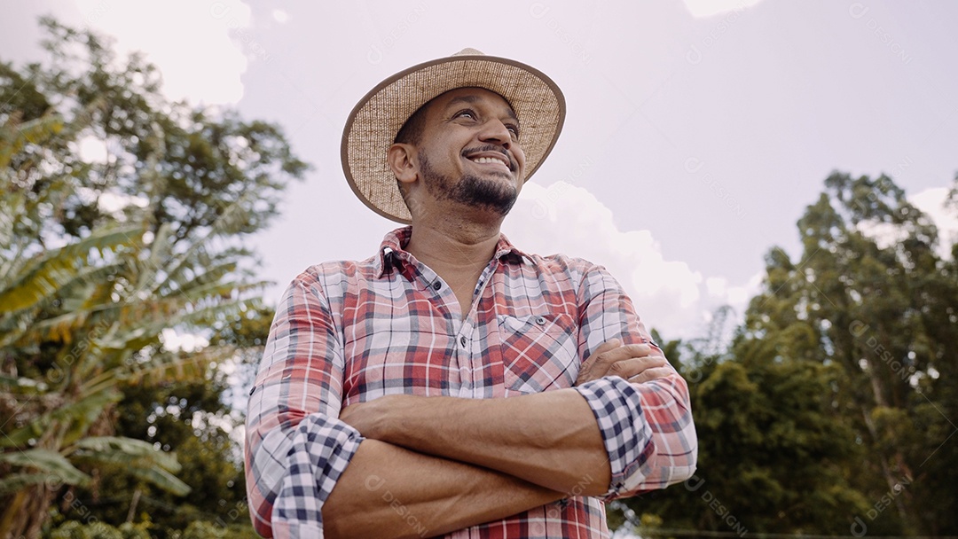 Retrato de jovem agricultor com mãos cruzadas na camisa casual e chapéu na fazenda