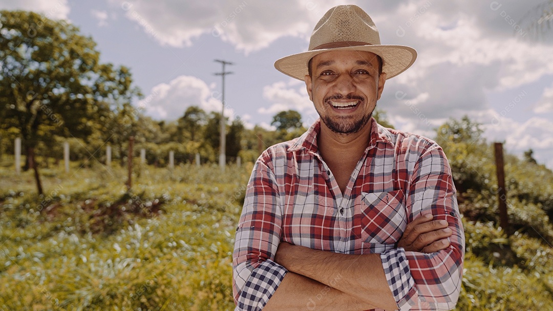 Retrato de jovem agricultor com mãos cruzadas na camisa casual e chapéu na fazenda