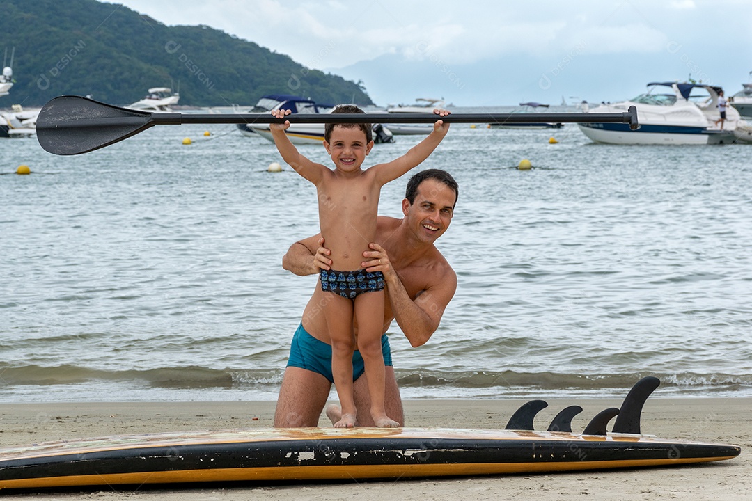 Pai sorridente segurando o filho na prancha de surf em pé.