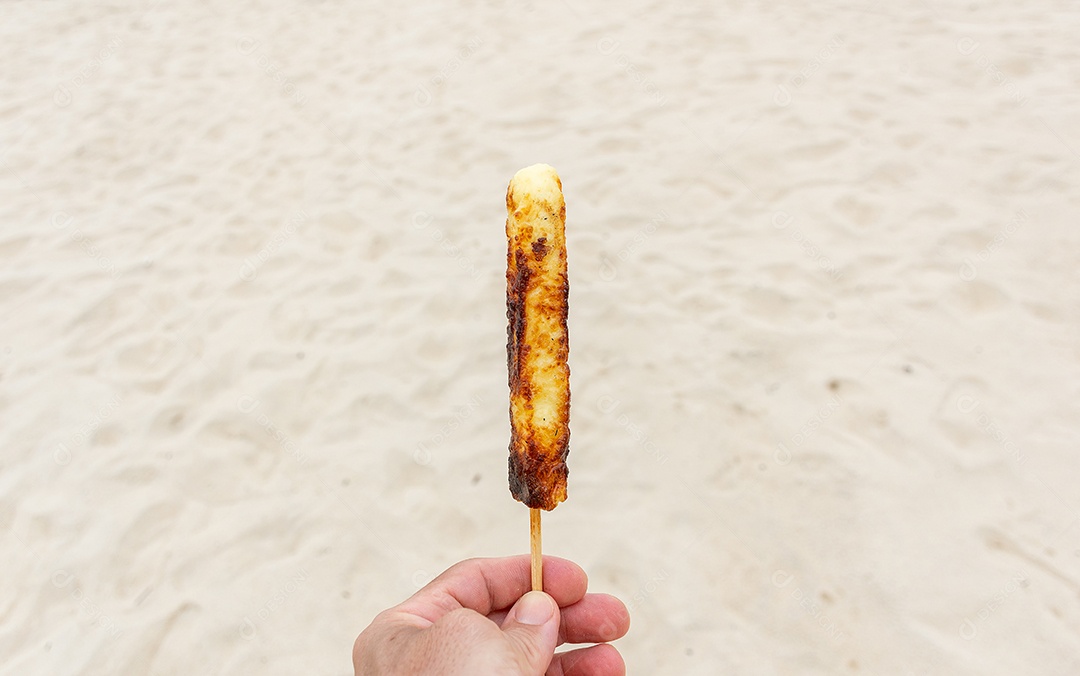 Requeijão assado preso a palito areia da praia ao fundo