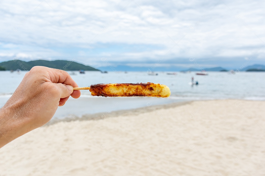 Requeijão assado preso a palito areia da praia ao fundo