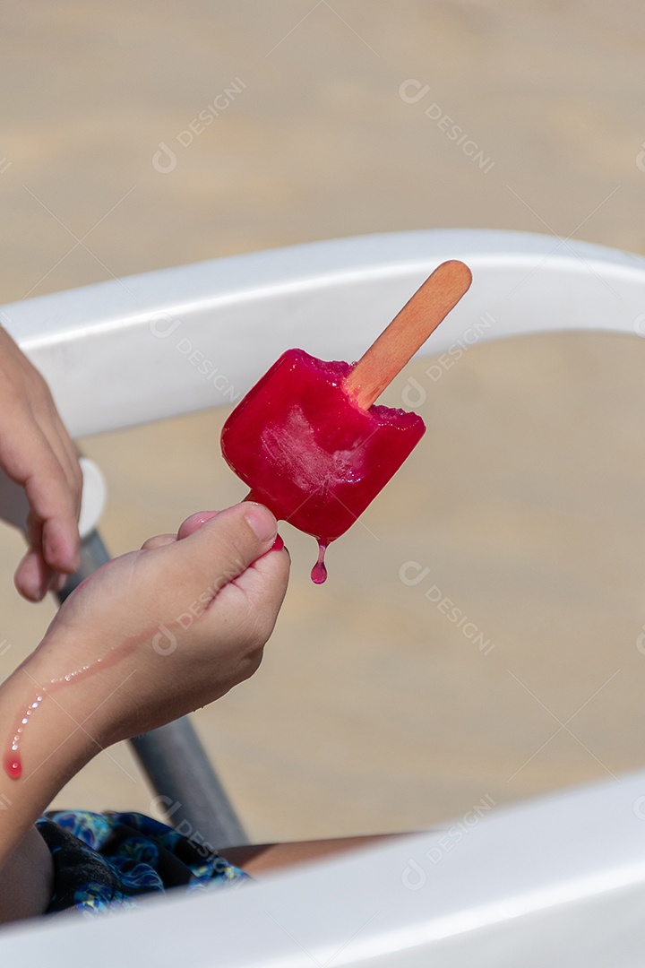 Criança com picolé de morango na praia derretendo na mão