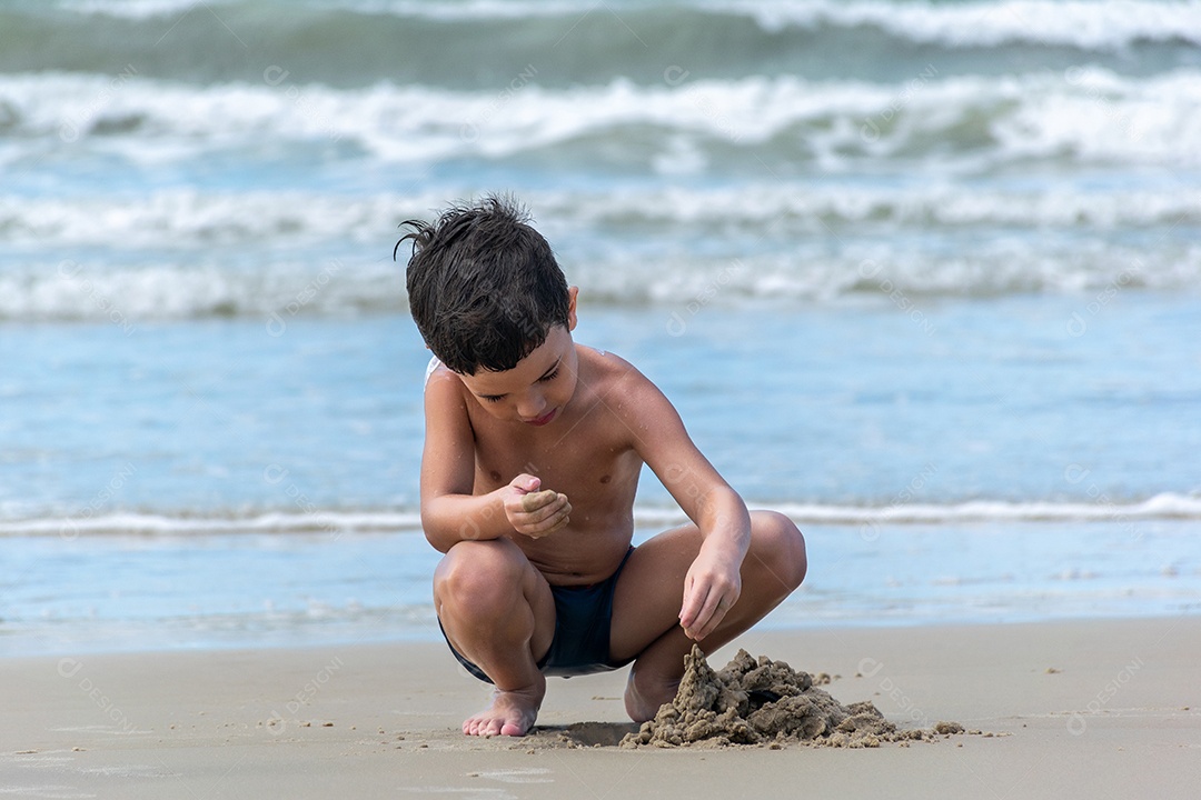 Criança sentada na areia da praia com os pés enterrados com as falésias da praia do Cotovelo ao fundo.