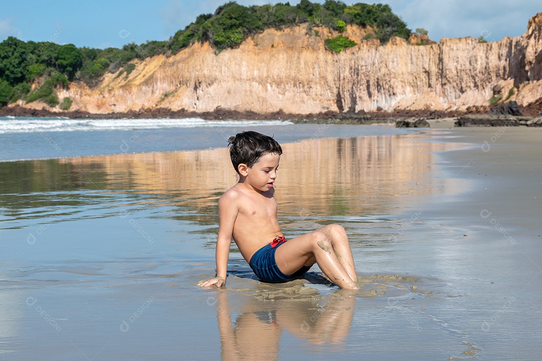 Criança sentada na areia da praia com os pés enterrados com as falésias da praia do Cotovelo ao fundo.