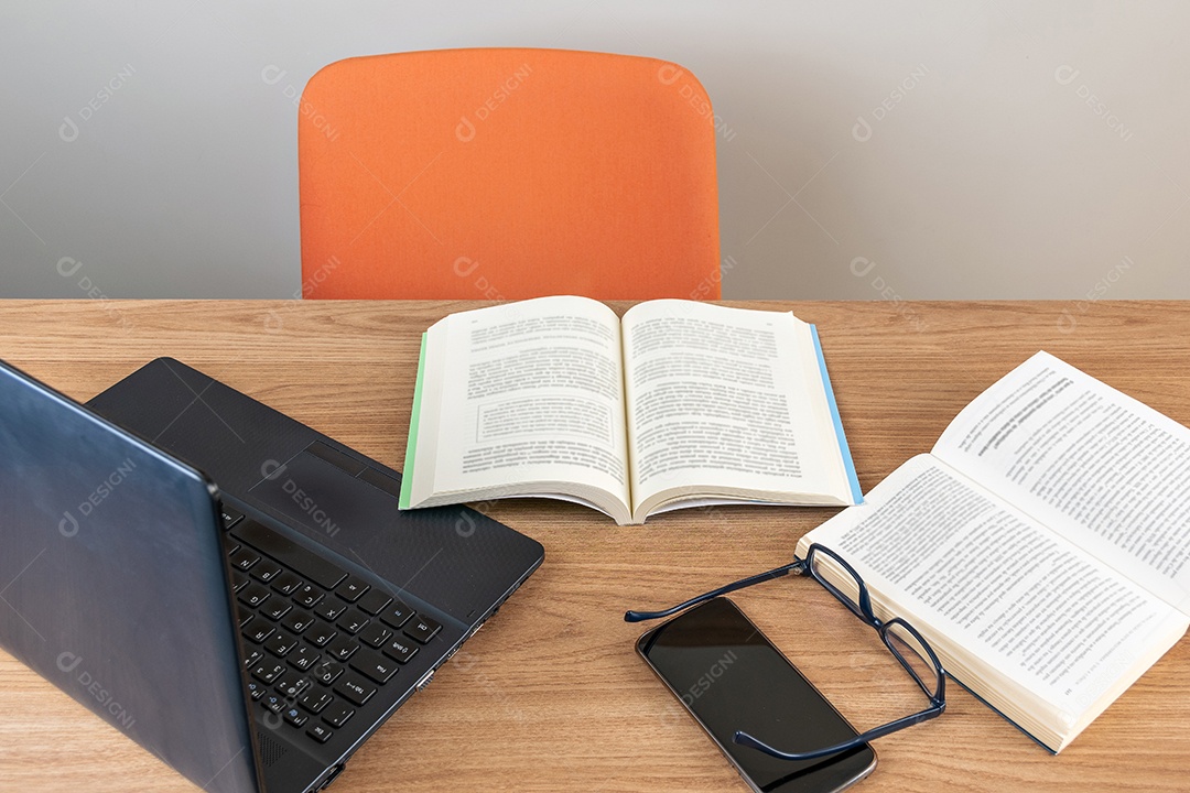 Mesa de madeira com cadeira laranja, livros abertos, celular e óculos