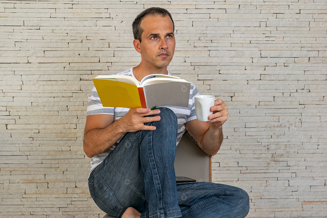 Homem de meia idade sentado na cadeira, lendo um livro, segurando uma xicara de café
