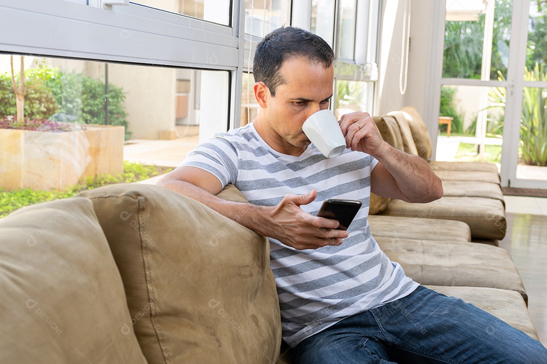 Homem de meia idade sentado no sofá, tomando café e usando o celular.