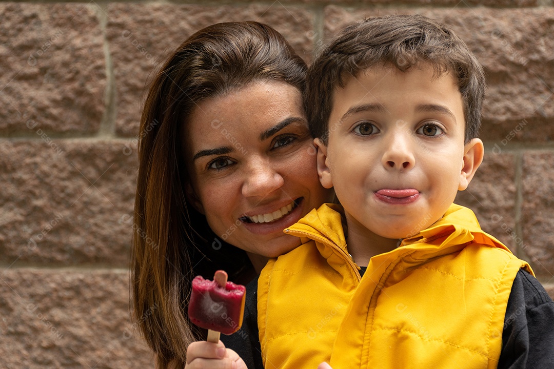 Mãe sorridente atrás de seu filho segurando um picolé com colete amarelo