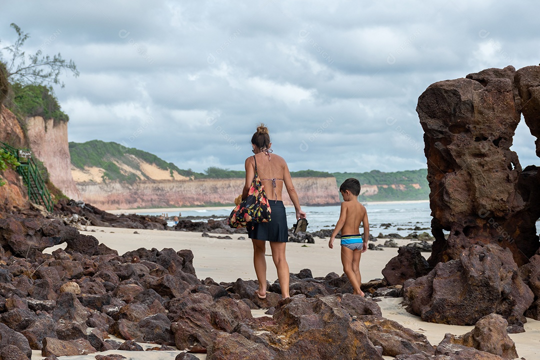 Mãe e filho de costas caminhando entre as pedras, indo para praia