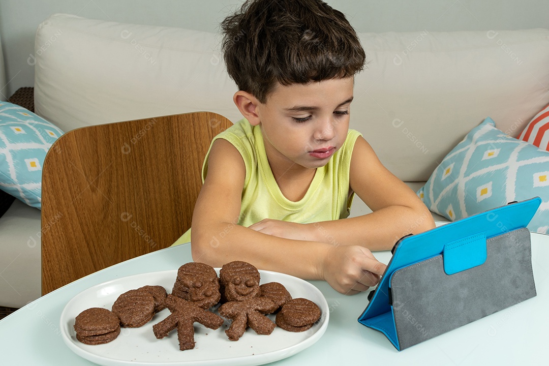 Criança em quarentena, comendo biscoitos e mexendo no tablet.