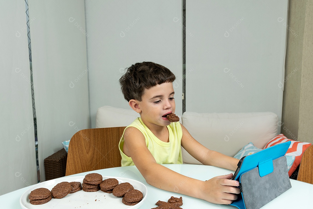 Criança em quarentena, comendo biscoitos e mexendo no tablet.