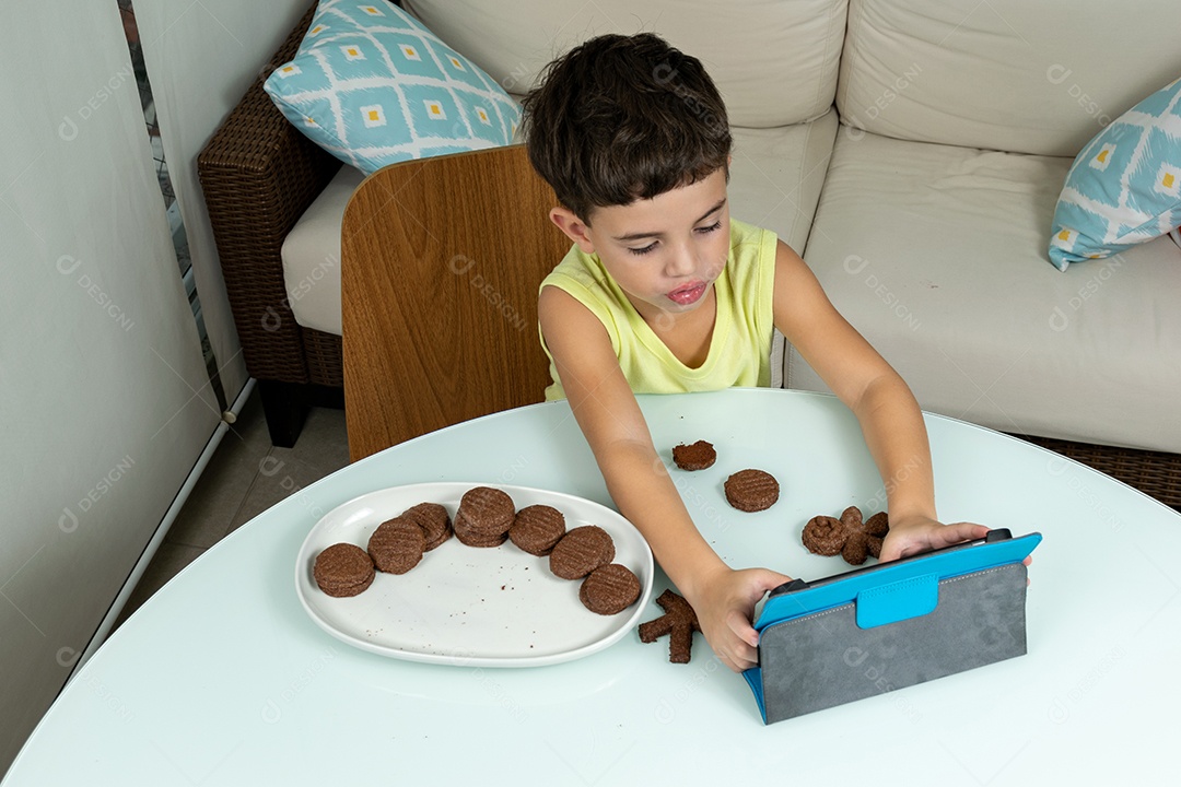 Child in quarantine, eating cookies and playing with tablet.
