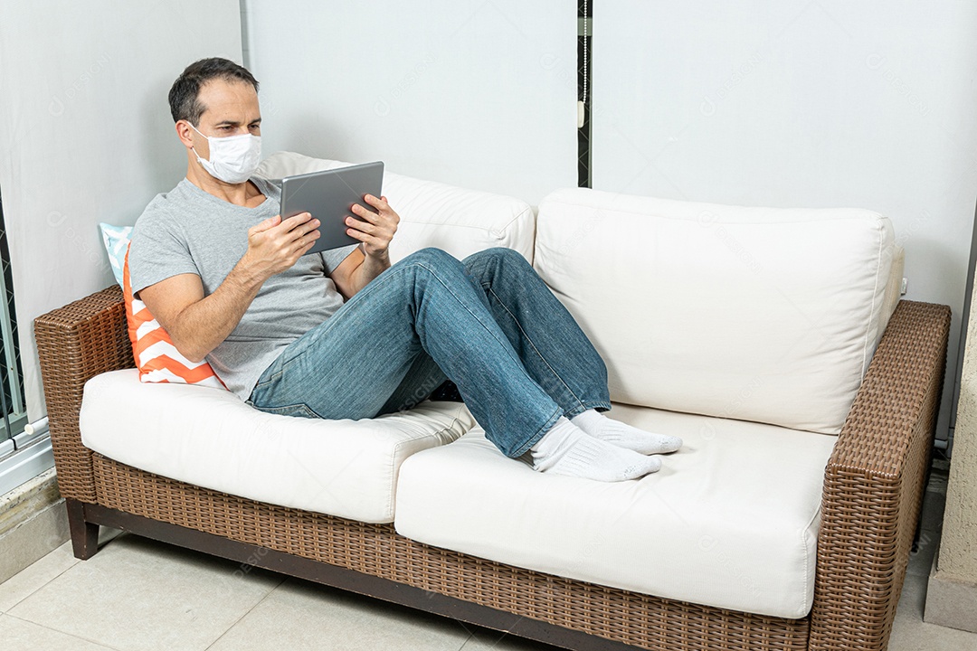 Homem de 44 anos com máscara assistindo ao tablet no sofá.