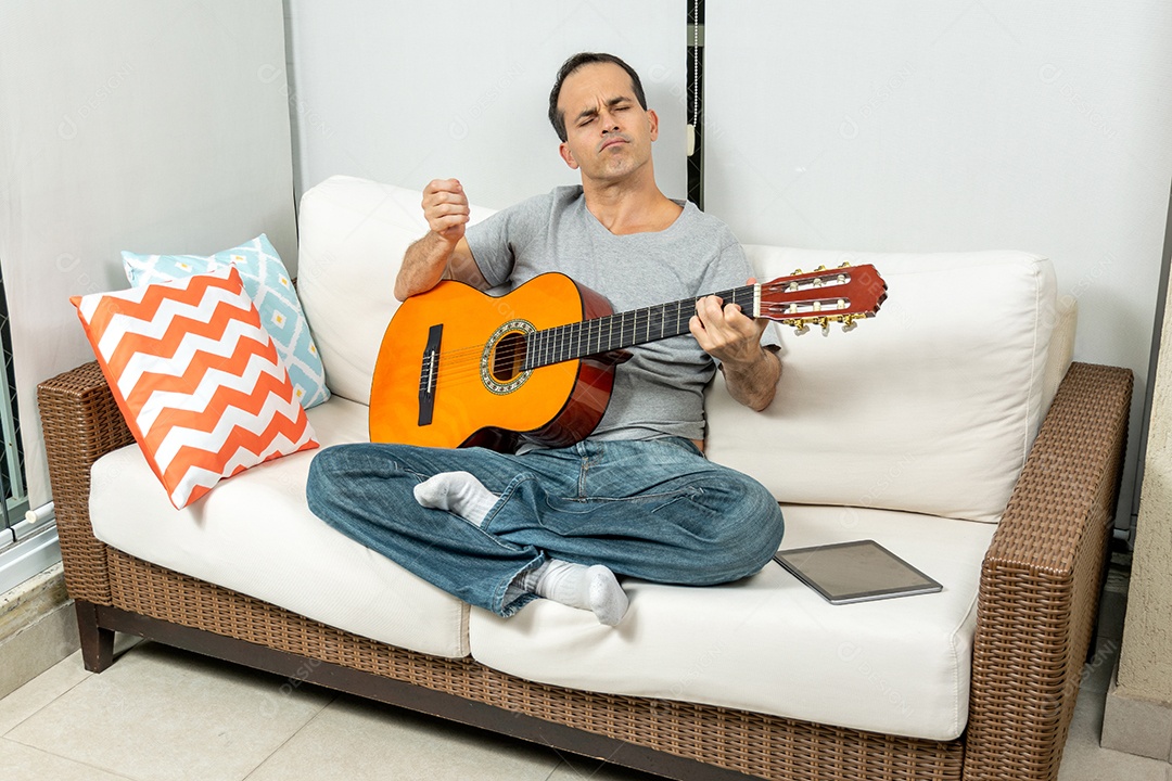 Homem maduro fazendo careta e tocando violão no sofá.