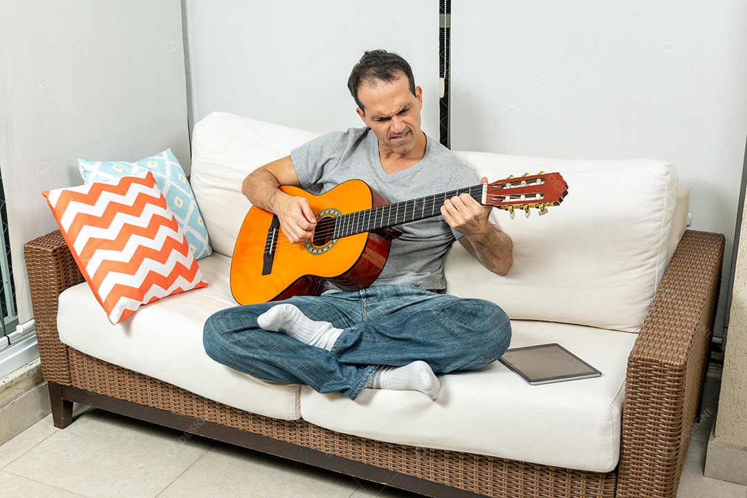 Homem maduro fazendo careta e tocando violão no sofá.