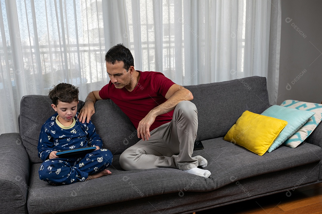 Homem jovem sentado sobre sofá com seu filho sobre conforto de sua casa