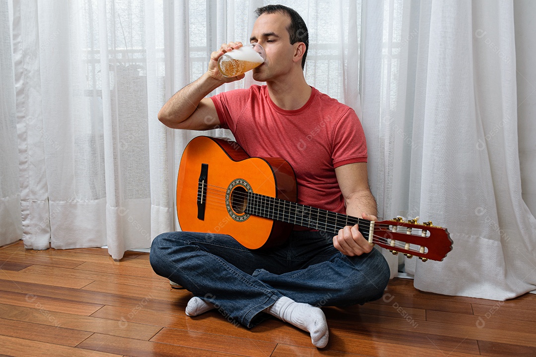 Homem maduro 44 anos sentado no chão e interagindo com um violão e um copo de cerveja.