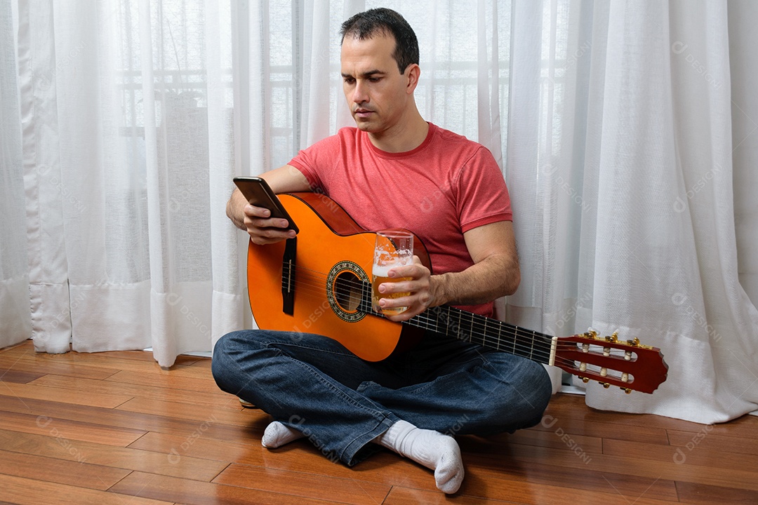 Homem maduro 44 anos sentado no chão e interagindo com um violão e um copo de cerveja.