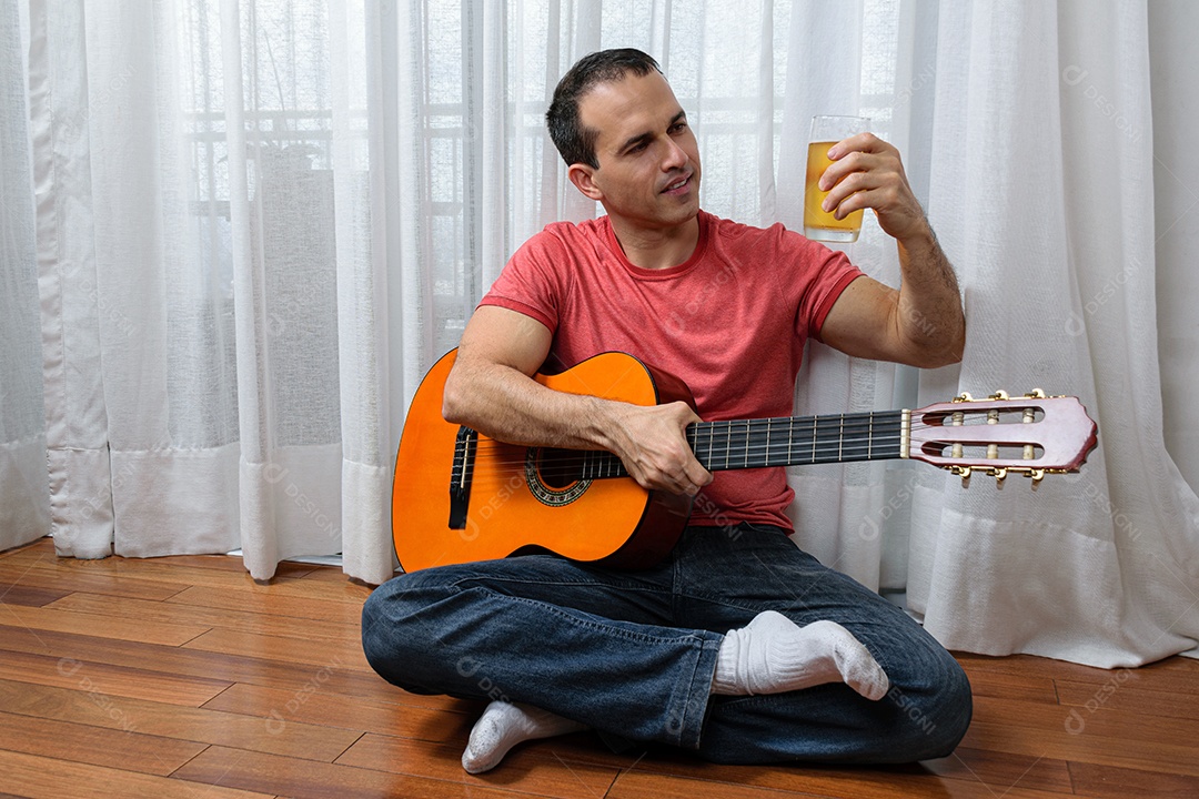 Homem maduro 44 anos sentado no chão e interagindo com um violão e um copo de cerveja.