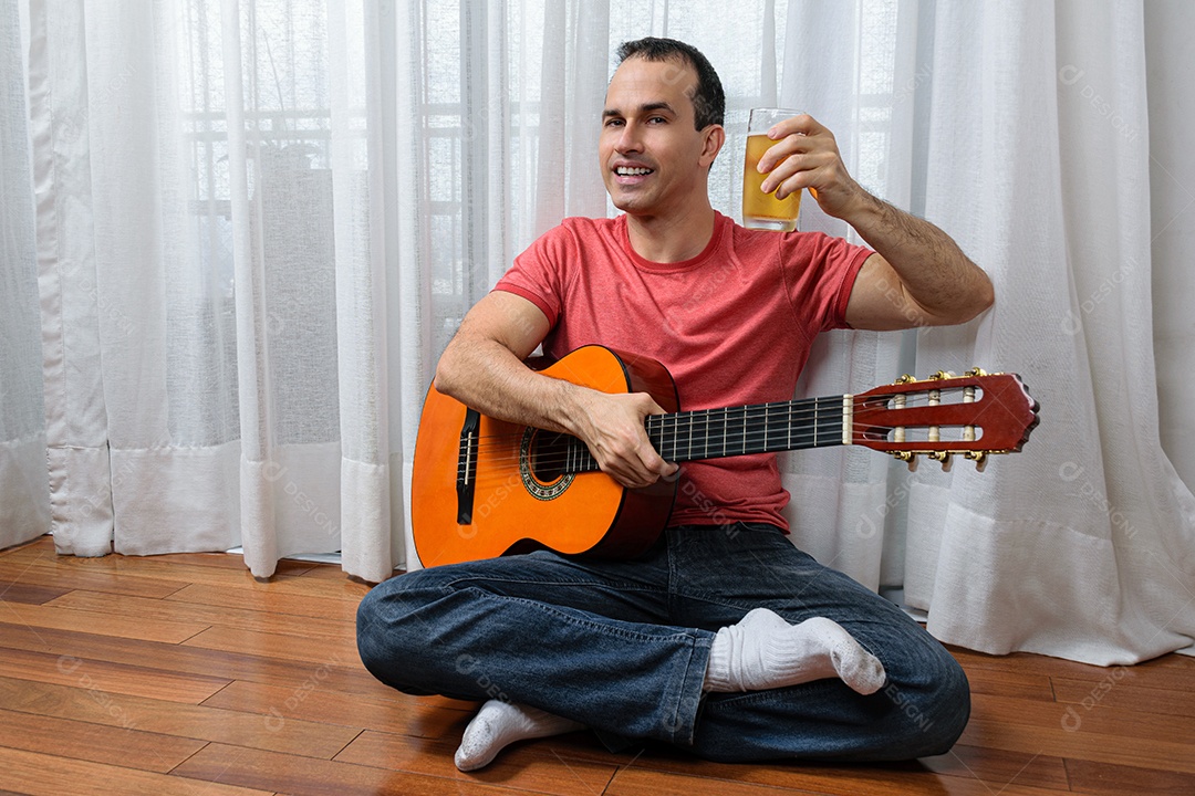 Homem maduro 44 anos sentado no chão e interagindo com um violão e um copo de cerveja.