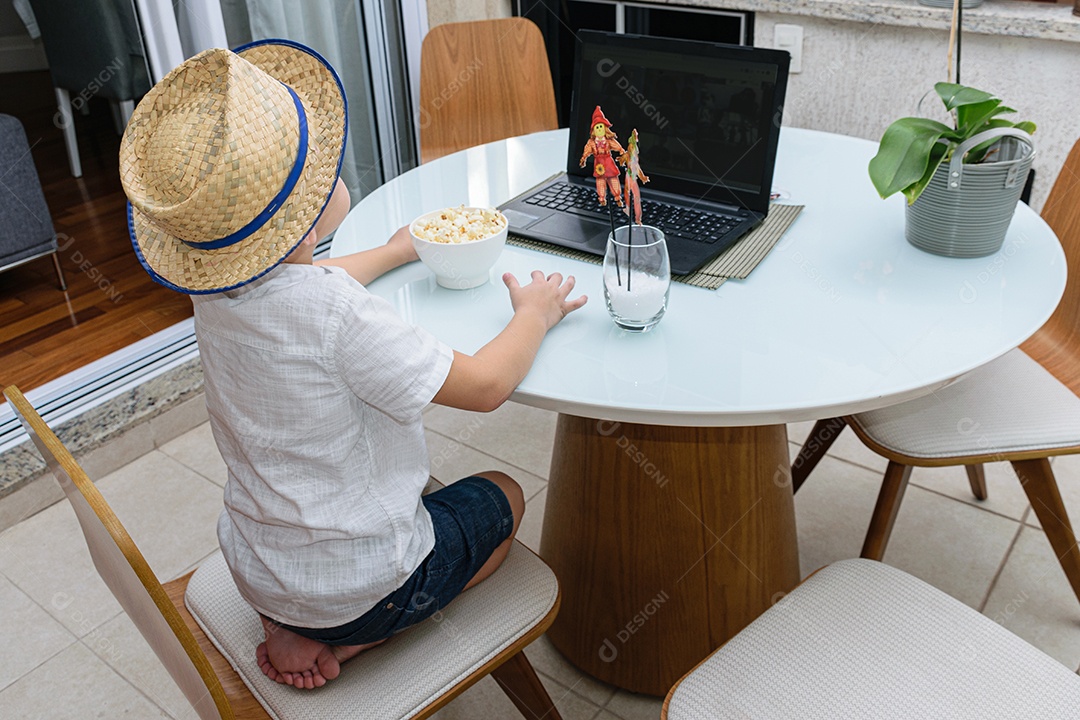 Criança de 7 anos com chapéu de palha e pipoca na frente do notebook