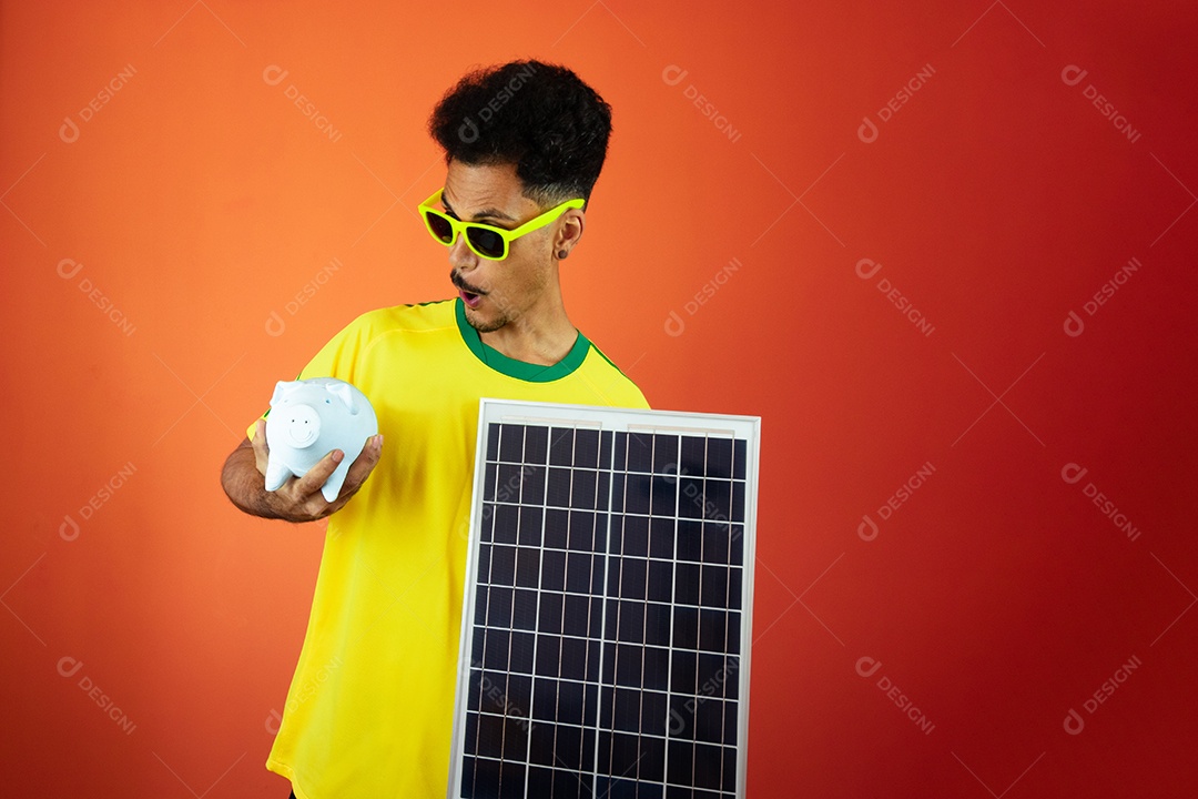 Jogador de futebol - homem negro comemorando segurando painel solar fotovoltaico e moeda de porco isolada em laranja