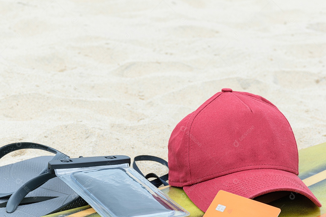 Smartphone e boné em uma prancha de surf, ao lado de chinelos na areia da praia.