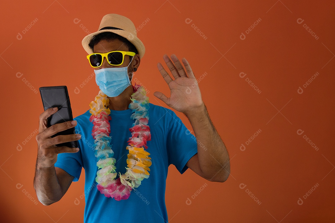 Homem negro em fantasia de carnaval e máscara pandêmica segurando um celular isolado em fundo laranja.