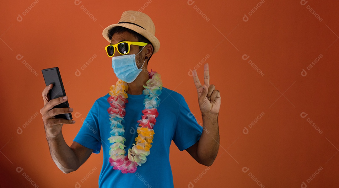 Homem negro em fantasia de carnaval e máscara pandêmica segurando um celular isolado em fundo laranja.