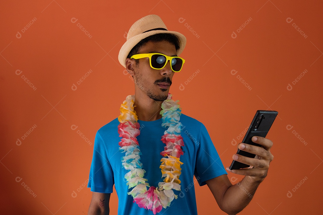 Homem negro em fantasia de carnaval e máscara pandêmica segurando um celular isolado em fundo laranja.