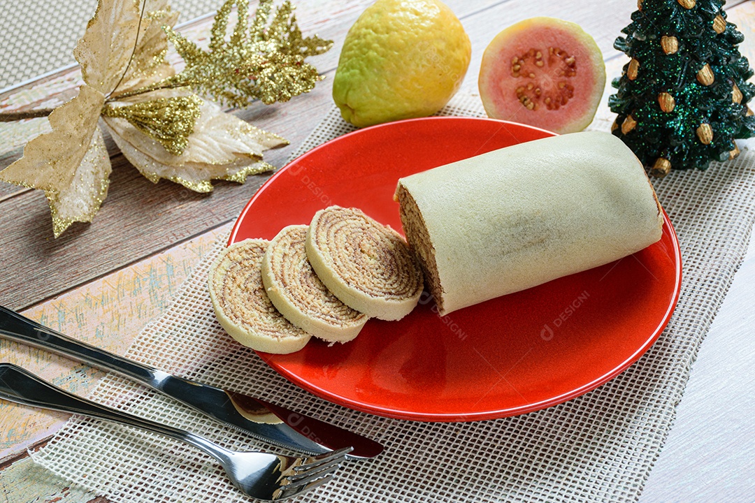Bolo de rolo fatiado em prato vermelho ao lado de goiabas.