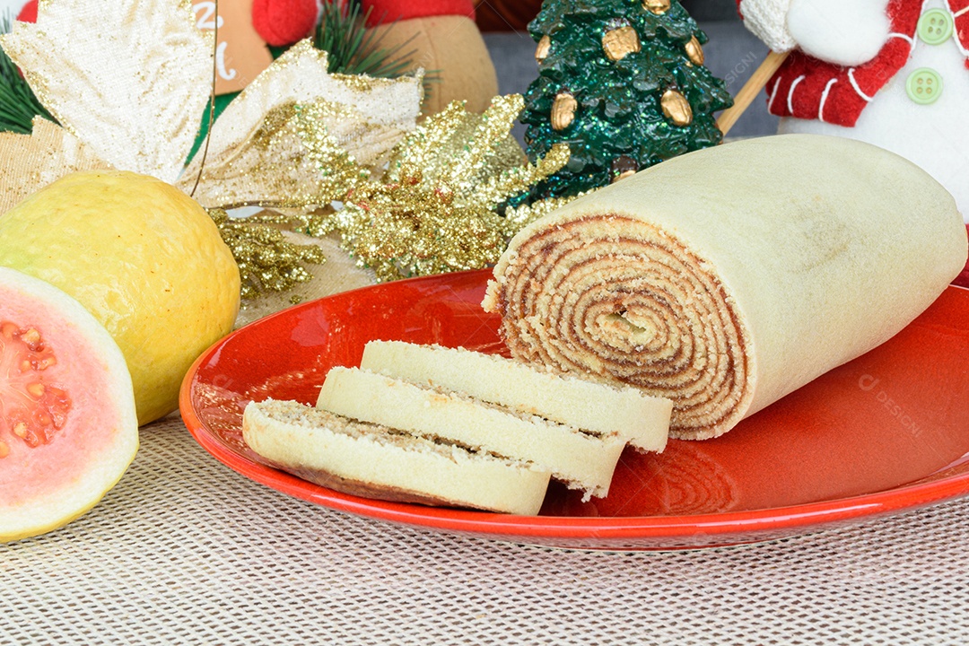 Bolo de rolo fatiado em prato vermelho ao lado de goiabas.