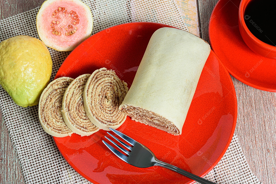 Bolo de rolo fatiado em prato vermelho ao lado de goiabas.