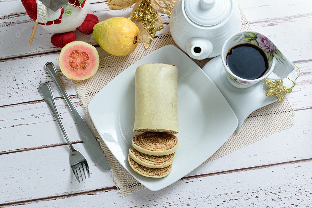 Bolo de rolo fatiado ao lado de talheres, xícara de café e goiabas, vista superior.