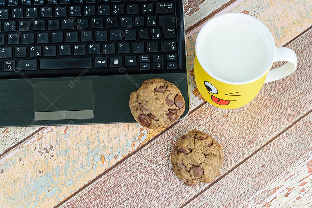 Biscoitos com gotas de chocolate no laptop, ao lado de uma caneca amarela com leite