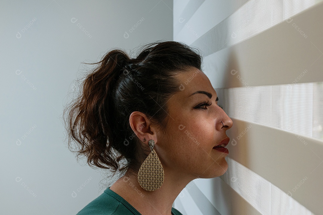 Mulher brasileira de 35 anos, com cara séria, olhando através da cortina