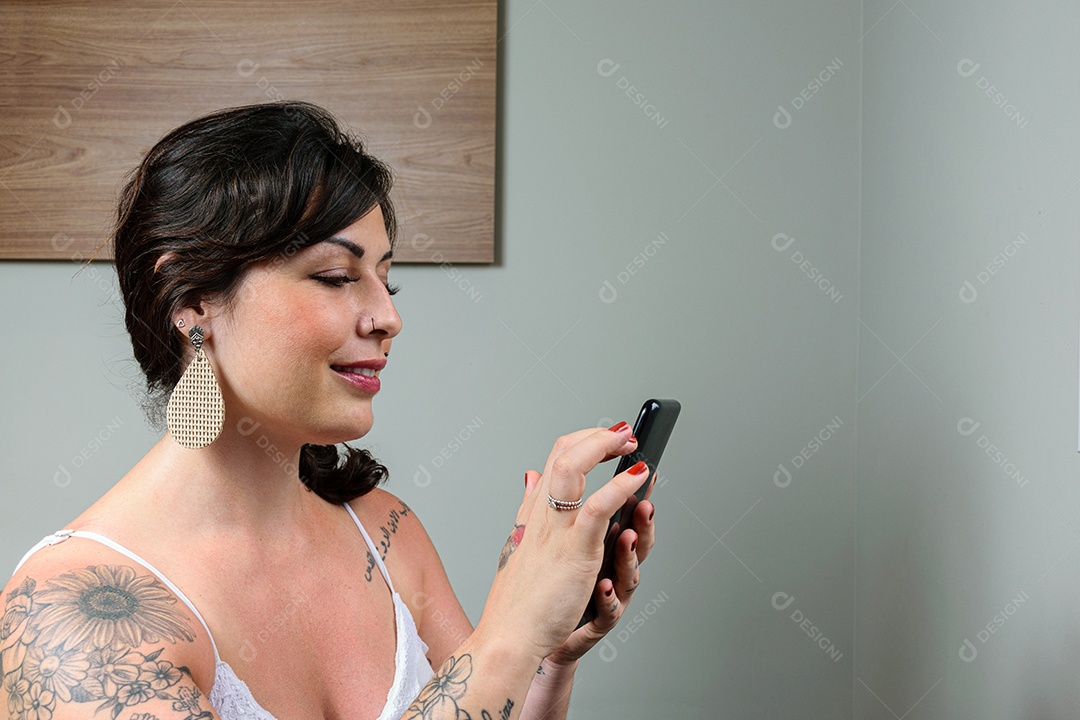Mulher brasileira, sorrindo, tatuada, segurando e interagindo com celular