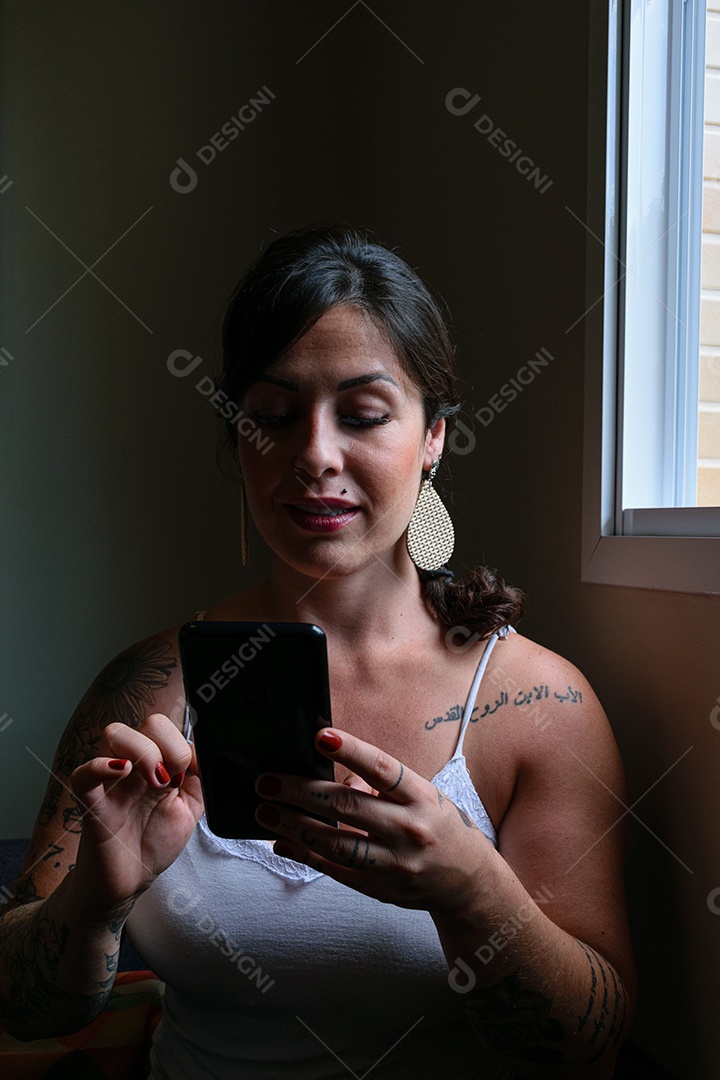 Mulher brasileira, tatuada, sub exposição, segurando e interagindo com seu smartphone